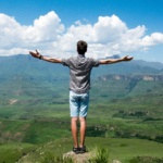 A man stands triumphantly with arms open, overlooking a scenic mountain range.