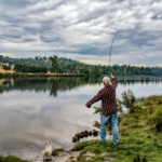 Elderly man fishing by a picturesque lake in Ouse, Tasmania, with a dog by his side.