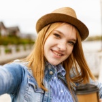 Young woman in a fedora hat takes a selfie with a coffee by the lake.