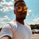 Young man in reflective sunglasses taking a selfie on a sunny day with clear skies and parked cars in the background.