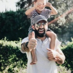 Father giving son a ride on shoulders, both laughing and smiling in a park setting.