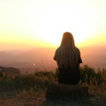 Woman sitting in nature, watching sunset over fields. Peaceful and serene outdoor scene.