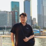 Teenage boy posing by a riverside with modern skyscrapers in the background.