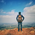 A man stands on a mountain edge appreciating the expansive scenic view below under a bright sky.