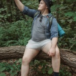 A hiker sitting on a tree trunk in the woods, capturing a selfie during a summer hike.